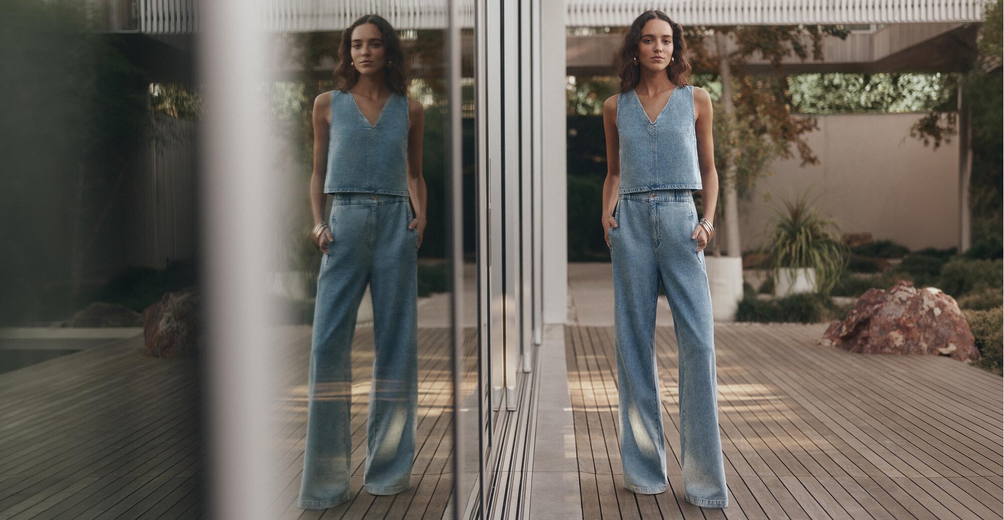 Woman wearing a sleeveless denim top and wide-leg jeans, standing beside a glass wall with a reflected mirror image.