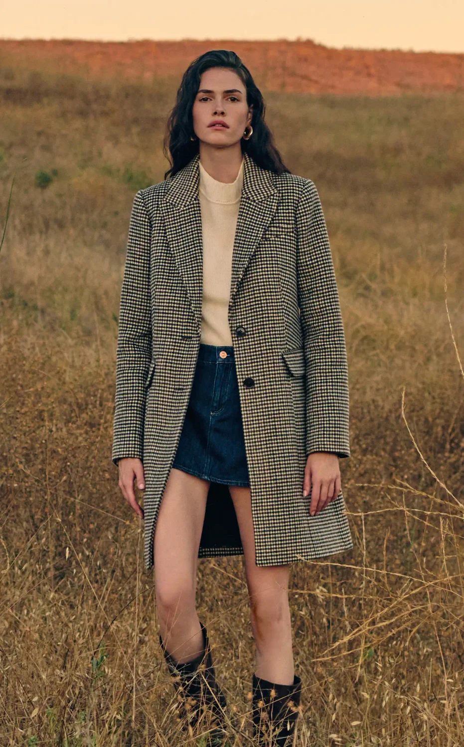 Woman standing in a field wearing a long houndstooth coat, an ivory sweater, denim mini skirt, and tall black boots