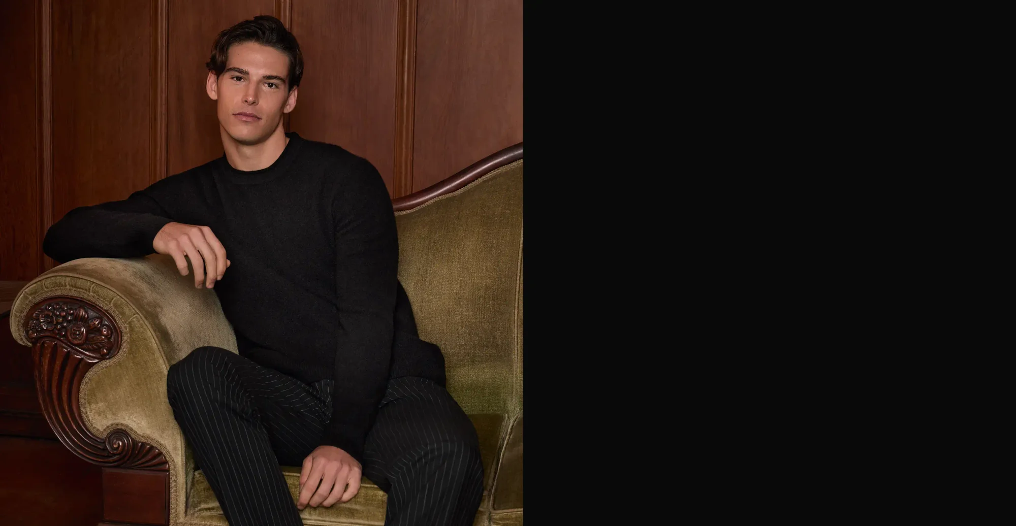 Man sitting on a couch wearing a black crewneck sweater, black pinstripe trousers and black sneakers