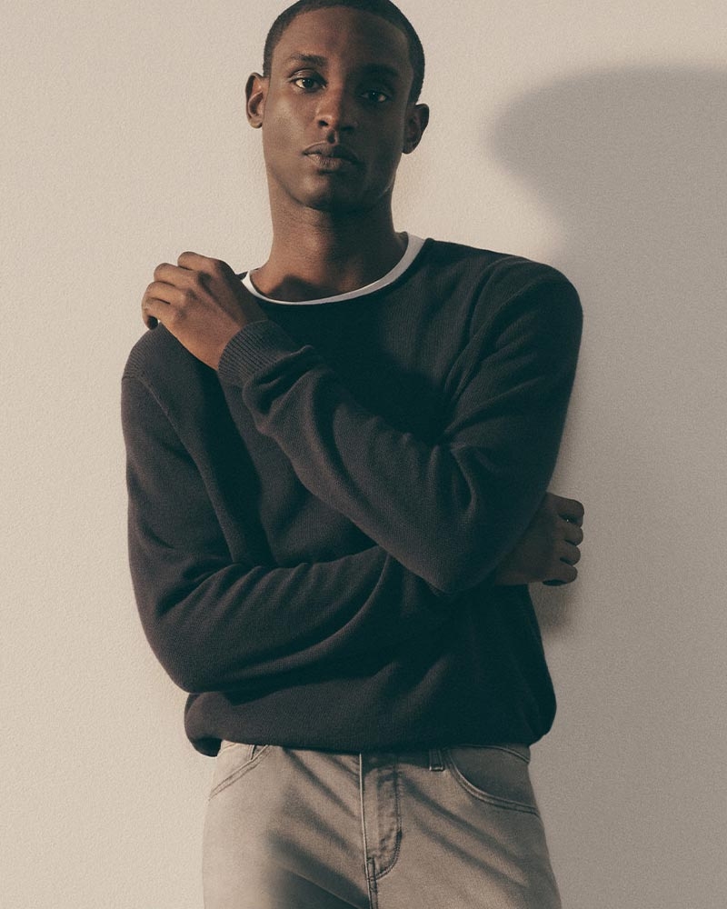 Man wearing a dark crewneck sweater and light gray jeans standing against a neutral wall.