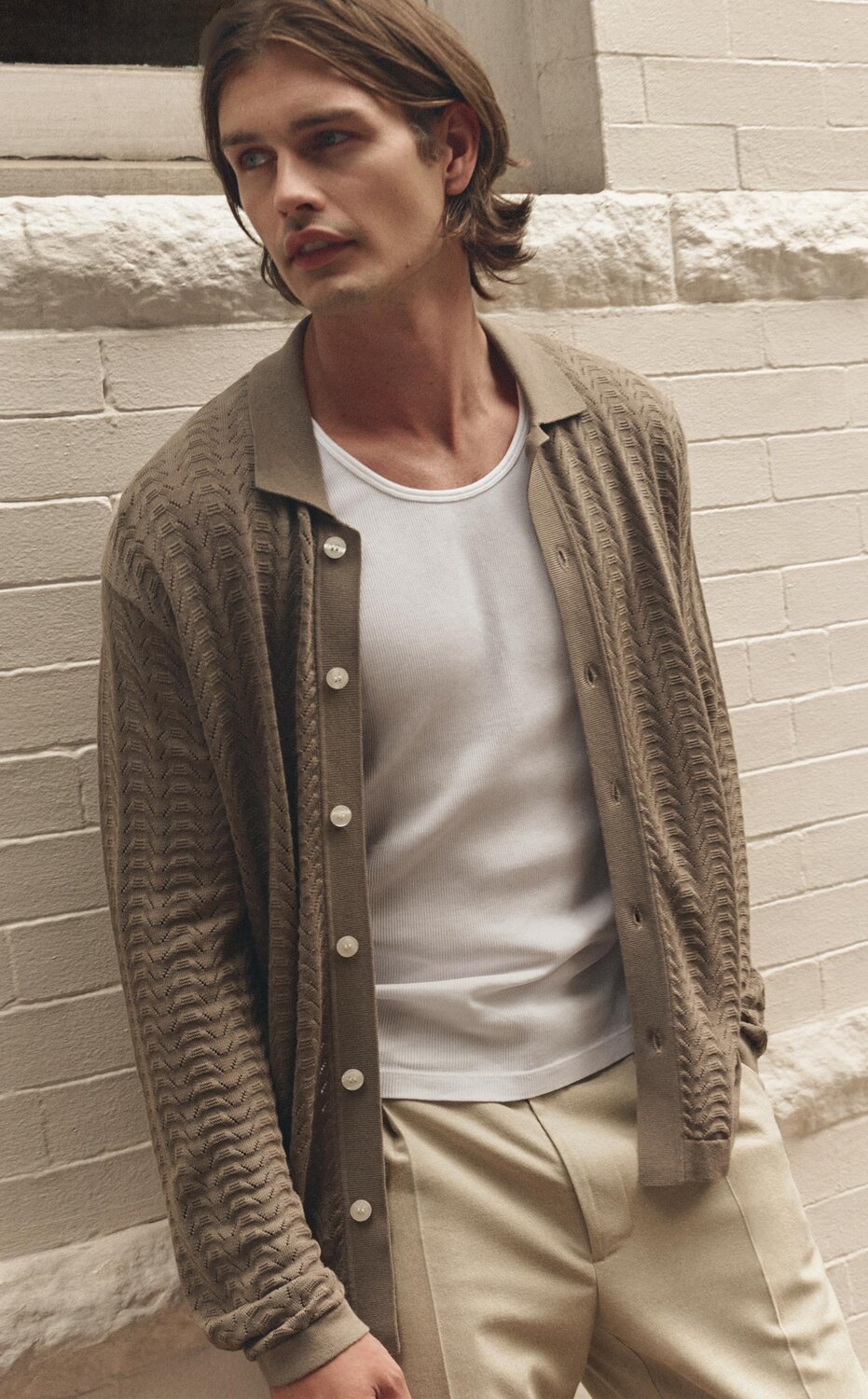 Man wearing a brown button-up sweater knit shirt over a white tank and beige trouser pants