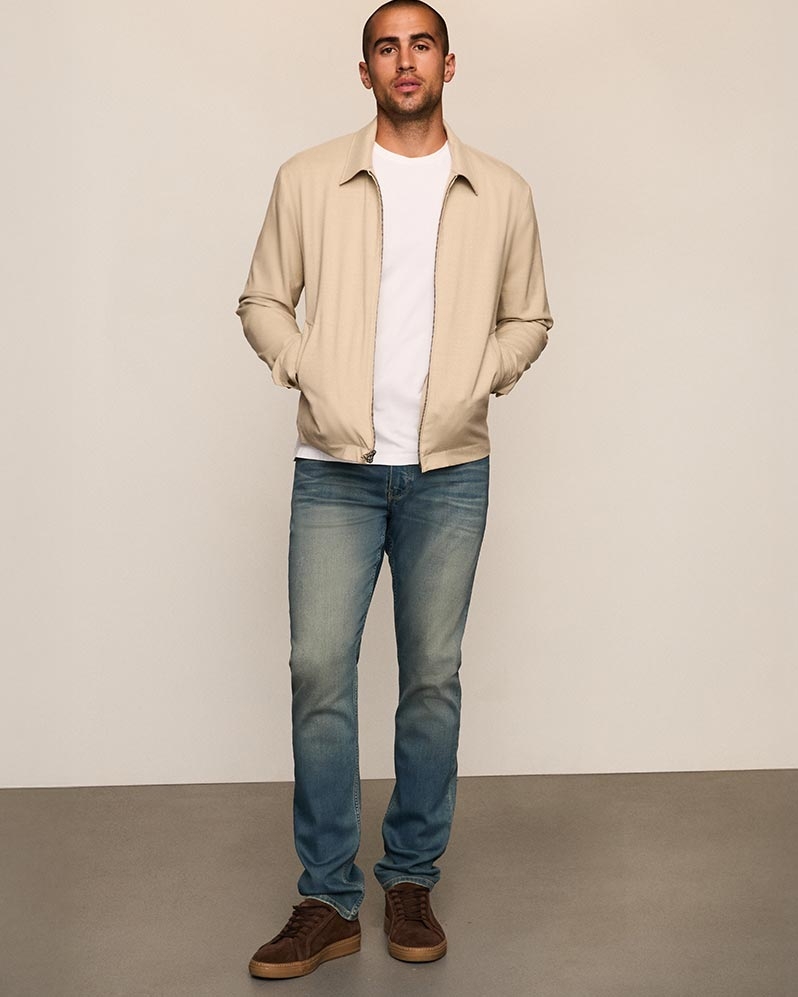 Man wearing a cream jacket over a white tee, dark wash jeans, and brown sneakers
