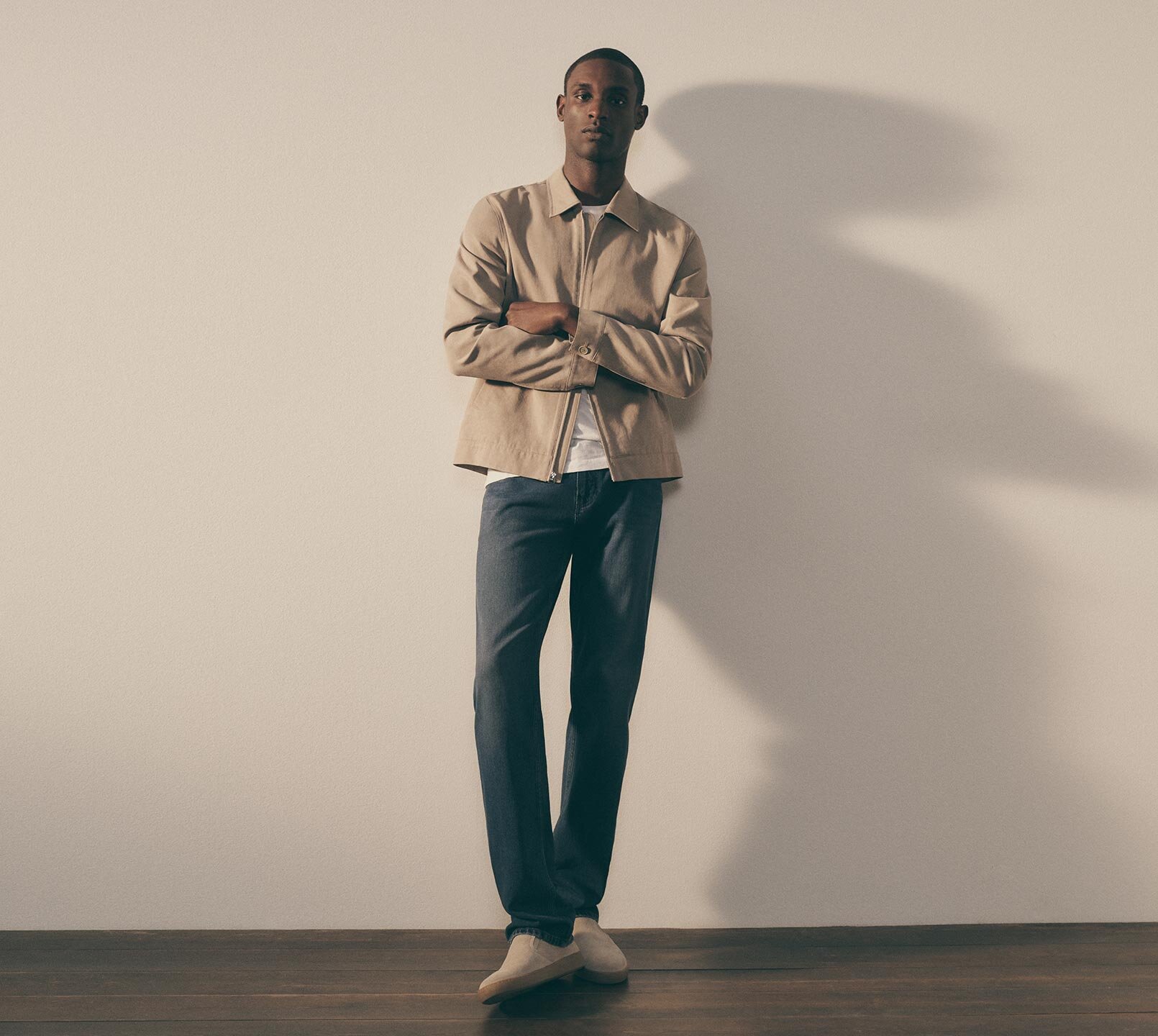 Man wearing a khaki jacket with a white t-shirt, medium wash blue jeans and beige slip on shoes all by PAIGE.