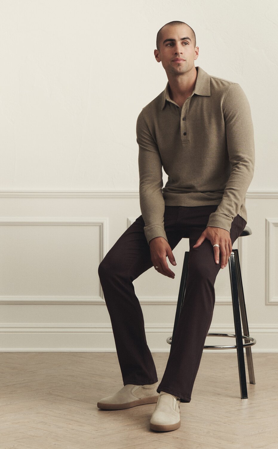 Man wearing a muted green long-sleeved polo knit blouse with a pair of dark brown straight-leg pants and beige suede slip-on sneakers, lounging on a stool against a cream wall with trimming and beige hardwood floors.