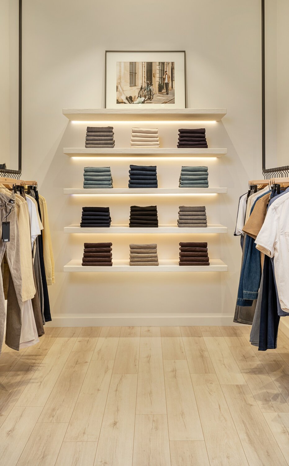 Interior of a PAIGE retail store featuring men's clothing racks, folded denim, and modern minimalist displays.