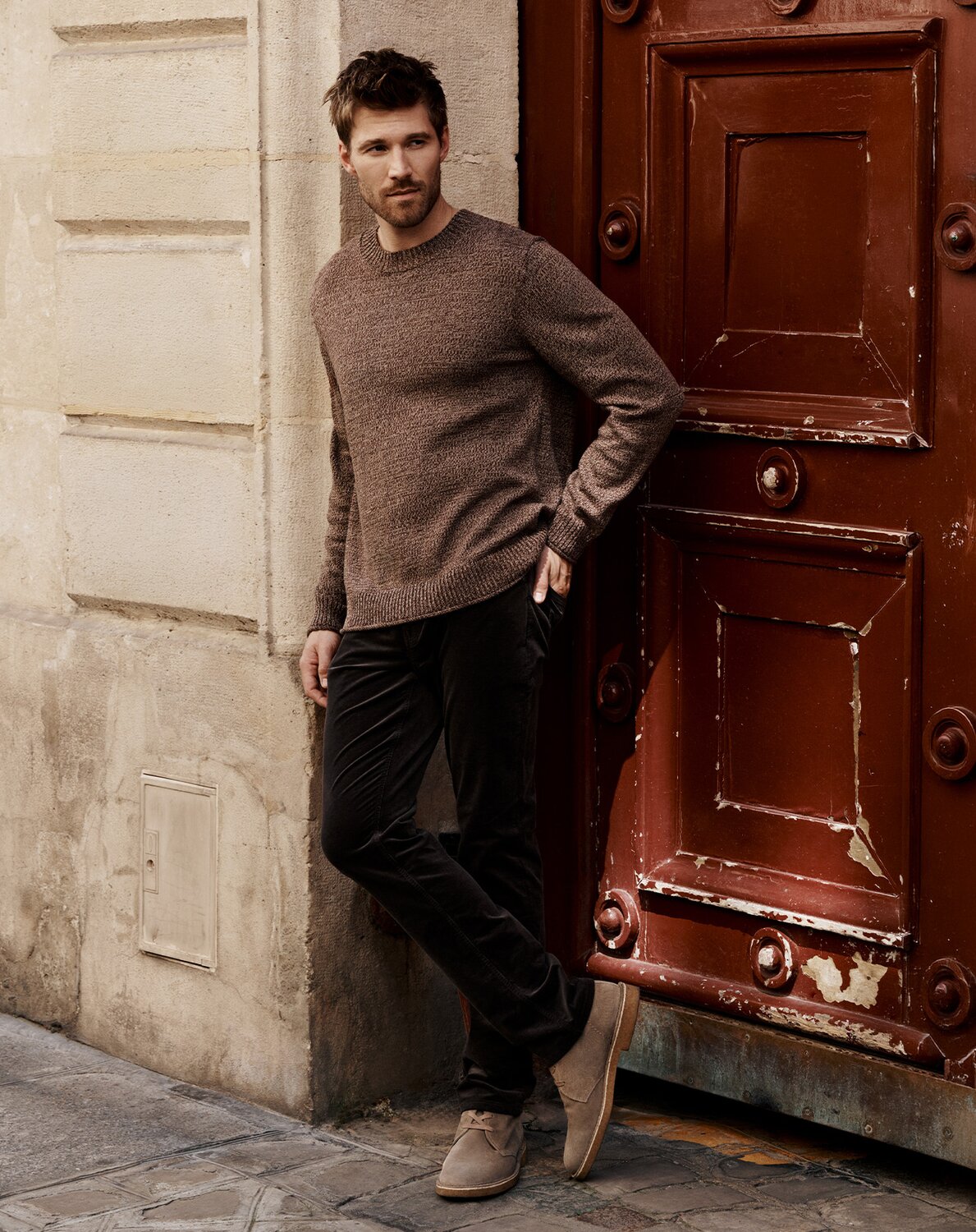 Man wearing a brown marl sweater and deep chocolate jeans with beige suede sneakers as he stands in front of a beige and red building exterior.