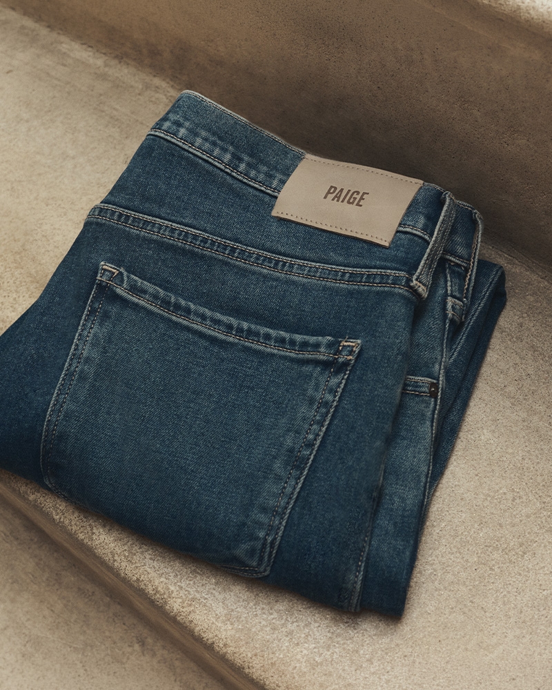 Folded pair of dark wash denim jeans with a back pocket and PAIGE label, placed on stone steps.