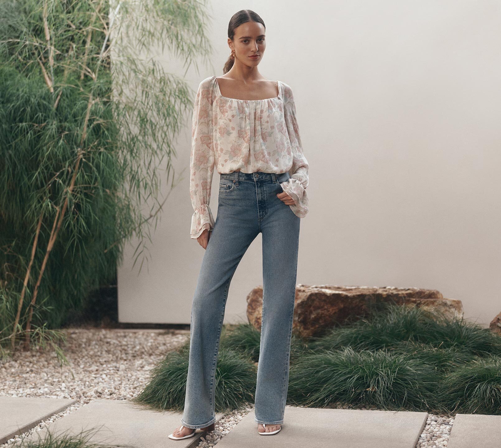 Woman wearing a floral print blouse with light wash blue jeans and white heeled sandals.
