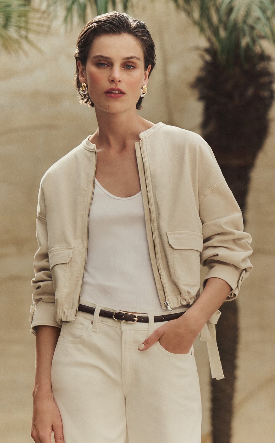 Female model wearing a cream-hued jacket with an adjustable tie waist and cargo-style pockets over a white scoop-neck tank and a pair of cream high-waist jeans fastened with a thin dark brown leather belt. She poses in front of a sunlit stone beige wall and trees.