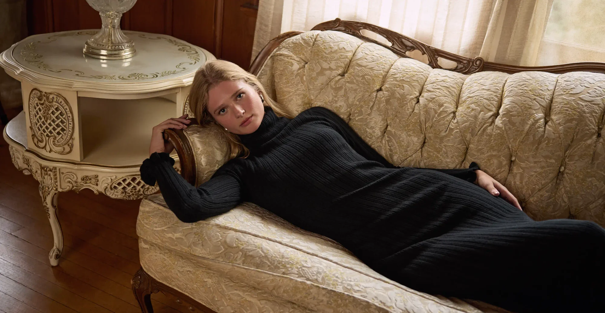 Woman laying on a couch wearing a long sleeve long black sweater dress and tall black boots