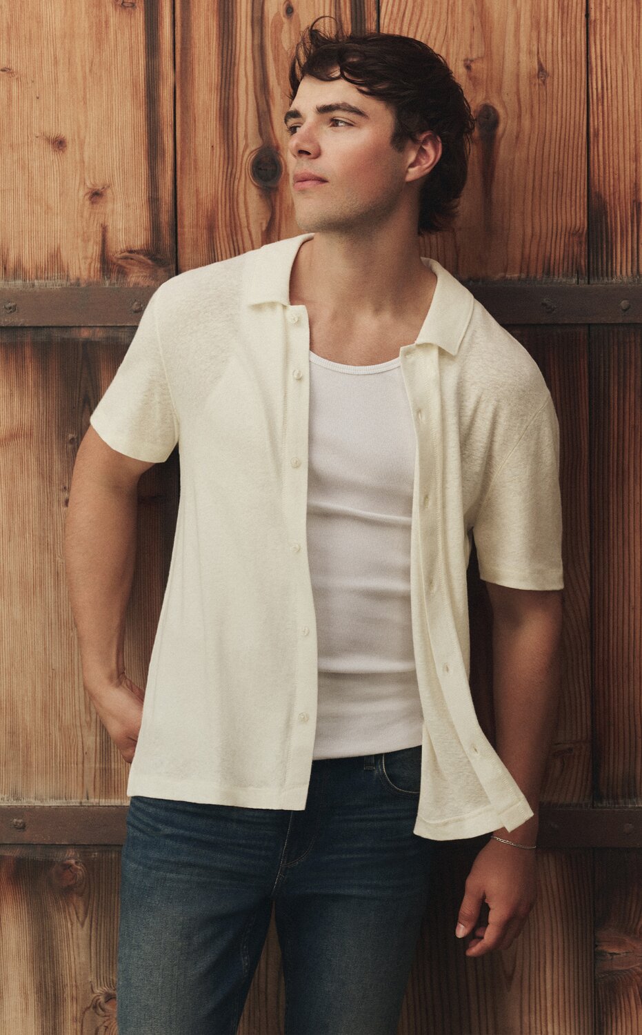 Male model wearing a white short-sleeve button-front shirt layered over a white tank top and dark-wash jeans, standing against a rustic wood-paneled wall.