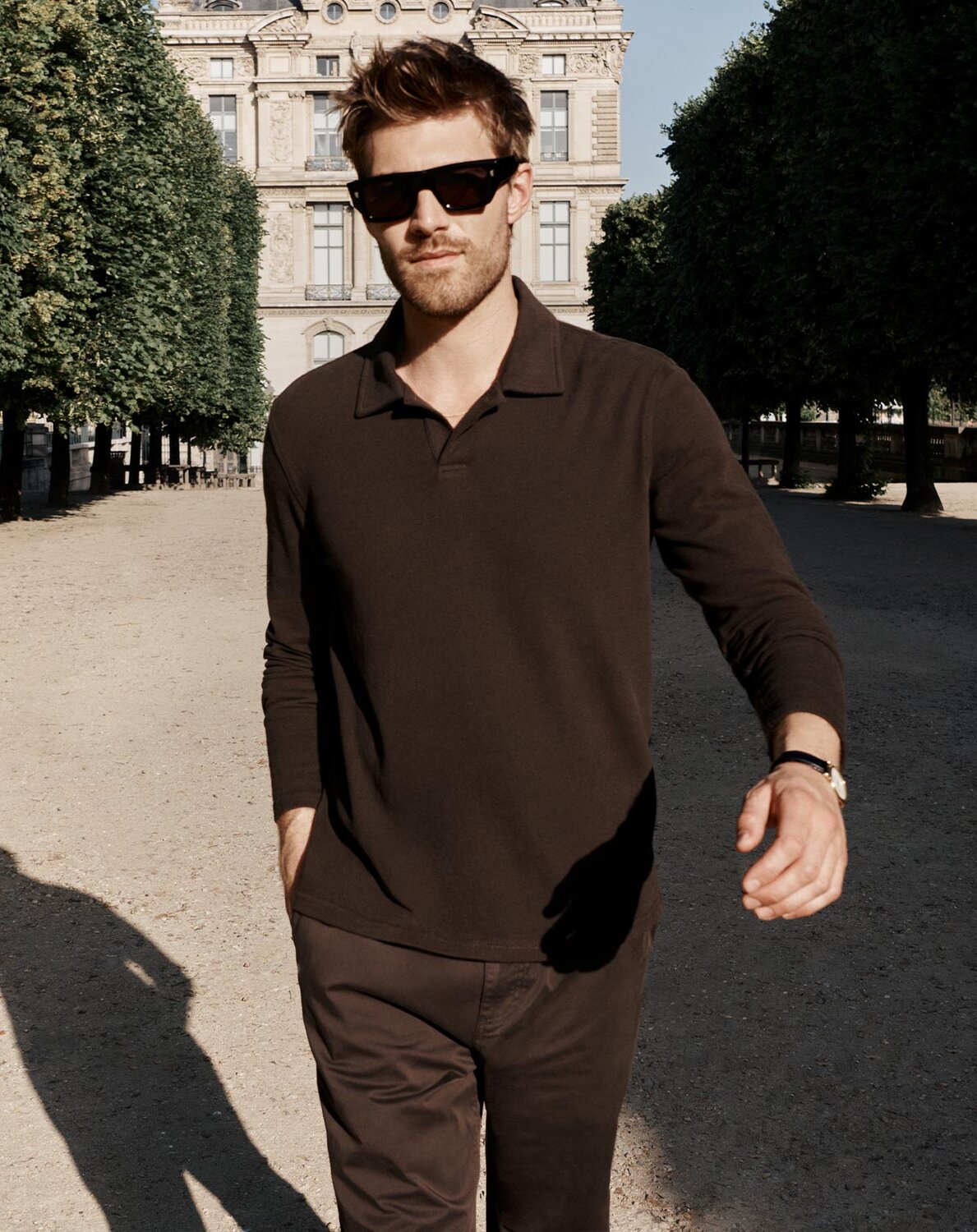 Man walking in a Parisian courtyard in a chocolate brown long-sleeved knit polo with a pair of brown corduroy pants and black sunglasses.