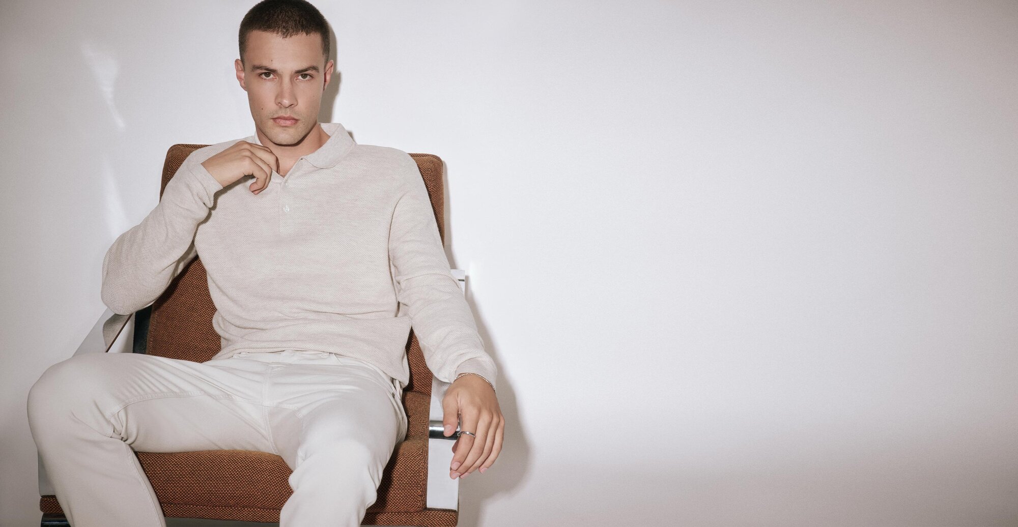 Man sitting in a chair wearing a cream long sleeve sweater polo and pale silver jeans