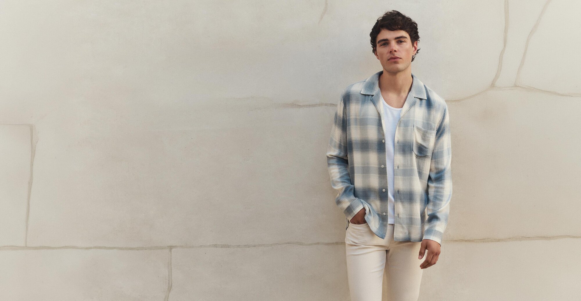 Male model wearing a light blue and cream plaid flannel long-sleeve button-up shirt over a white ribbed tank with light cream colored denim jeans, all by PAIGE, standing in front of a light cream colored wall.
