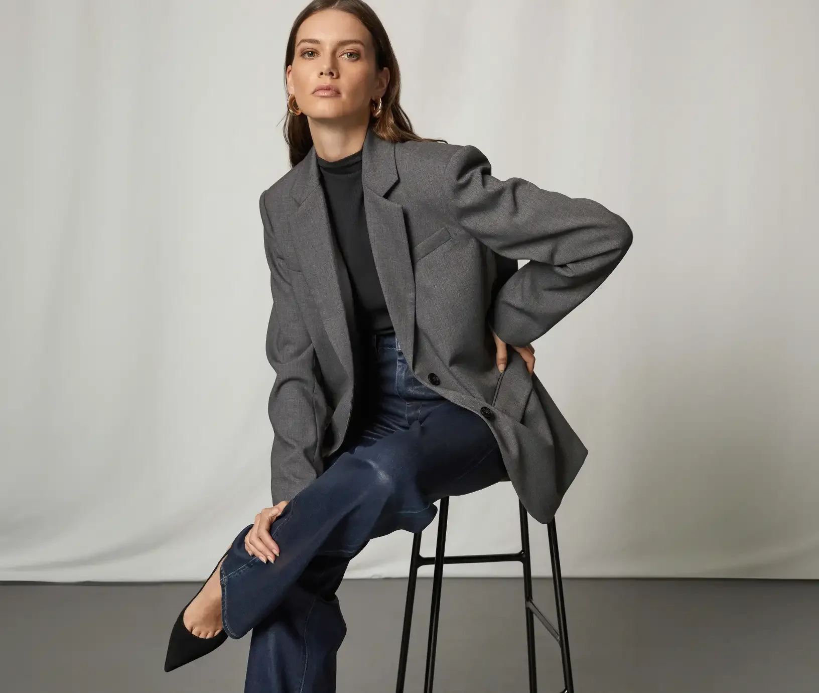 Woman sitting on a stool wearing an oversized grey blazer, black top, dark wash jeans and black pointed toe heels