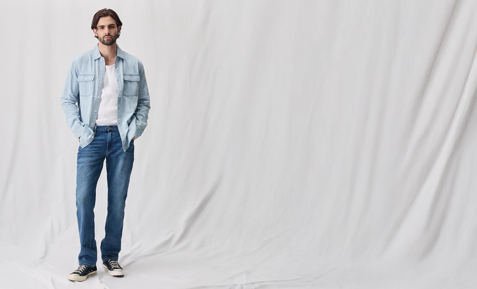 Man wearing a light wash denim shirt with a white tank top underneath, medium wash jeans and black sneakers