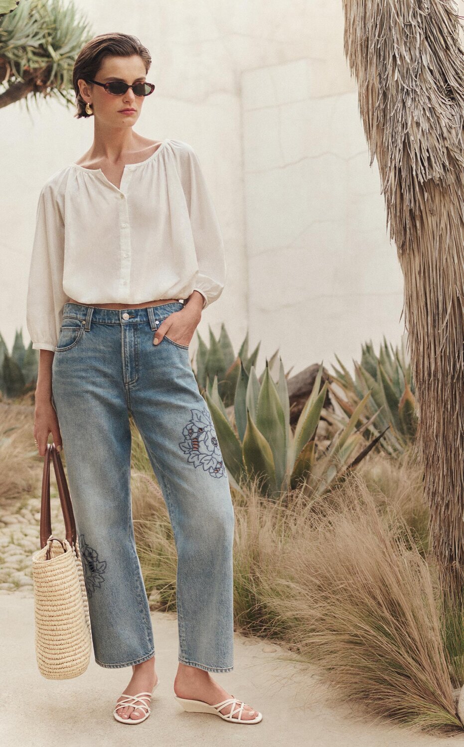 Model wearing a white button-front blouse and light-wash cropped jeans with floral embroidery, accessorized with sunglasses, woven tote, and white sandals, standing in a desert-inspired landscape with cacti, palms, and natural stone accents.