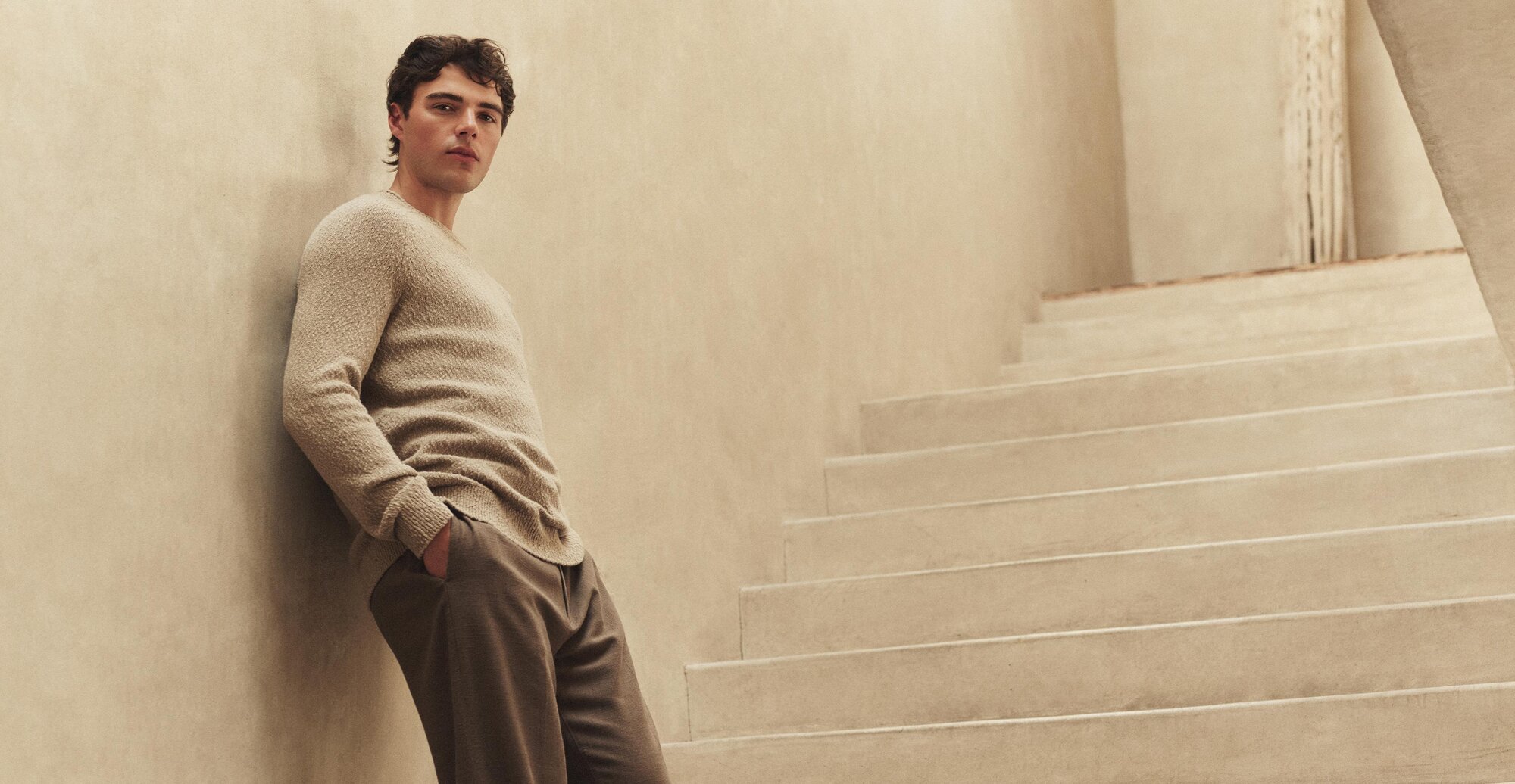 A man wearing a beige cotton sweater and brown linen pants leaning against a beige wall with beige steps leading up in the background.