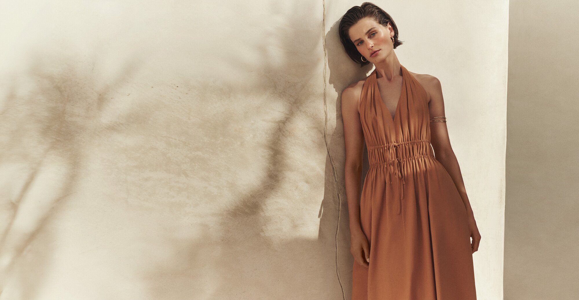 Model wearing an orange halter-neck ruched maxi dress, leaned against a sunlit beige wall.