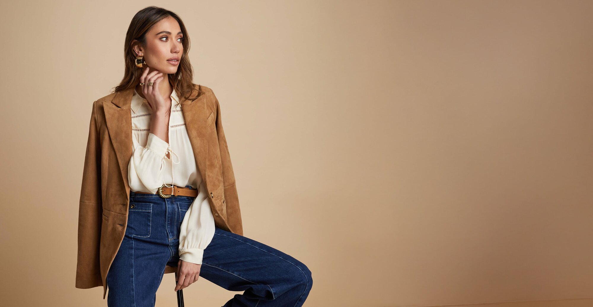 Woman sitting on stool wearing a brown suede blazer, white blouse, brown belt and dark wash jeans