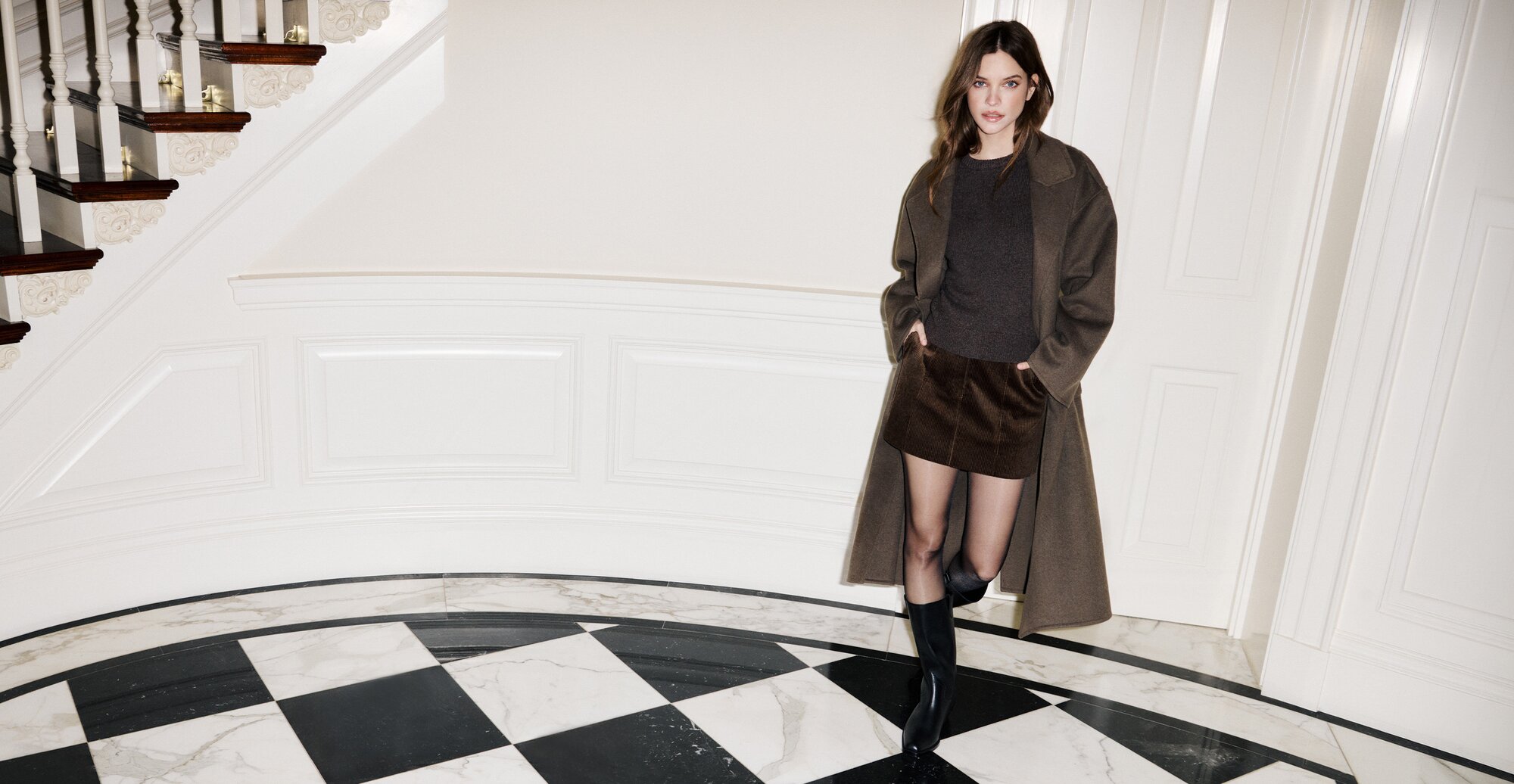 Barbara wearing a long dark taupe winter coat over a brown sweater and corduroy miniskirt with black leather boots, posed against a white door and marble checkerboard flooring.
