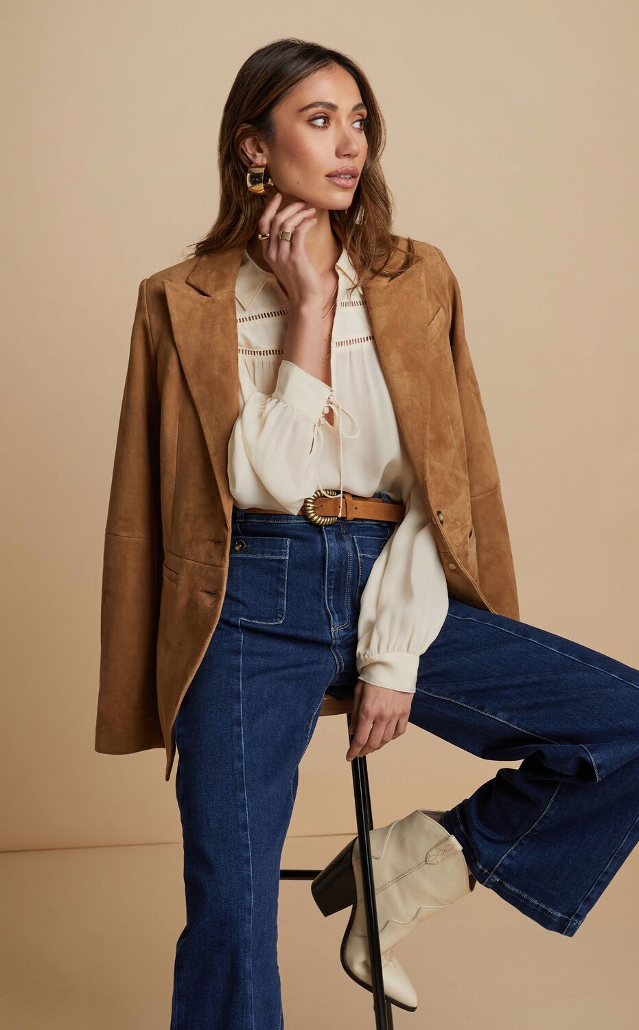 Woman sitting on stool wearing a brown suede blazer, white blouse, brown belt and dark wash jeans