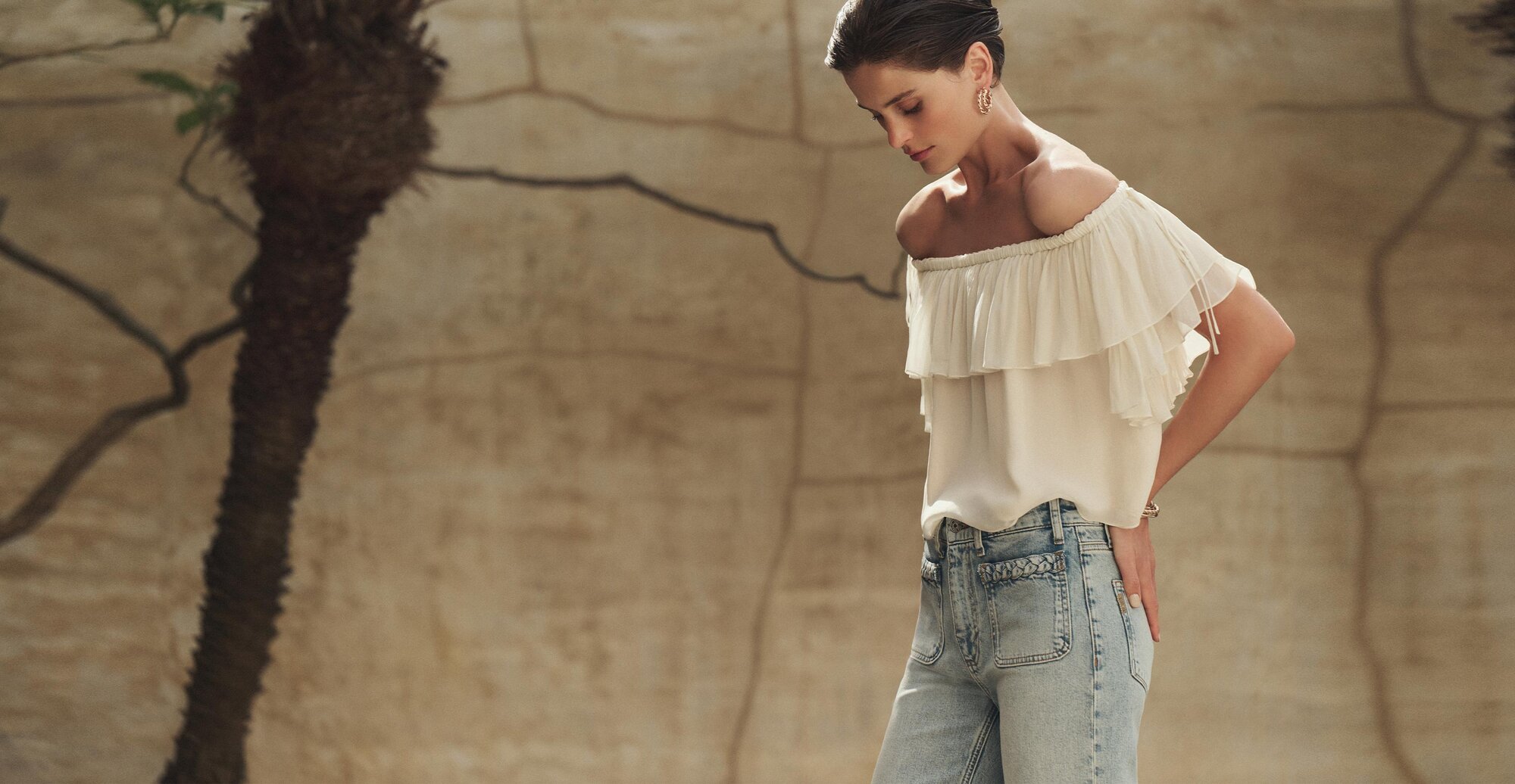 Woman wearing a white off-the-shoulder ruffle blouse and light-wash high-rise jeans with braided pocket detailing, standing against a sunlit stone wall with palm trees in the background.