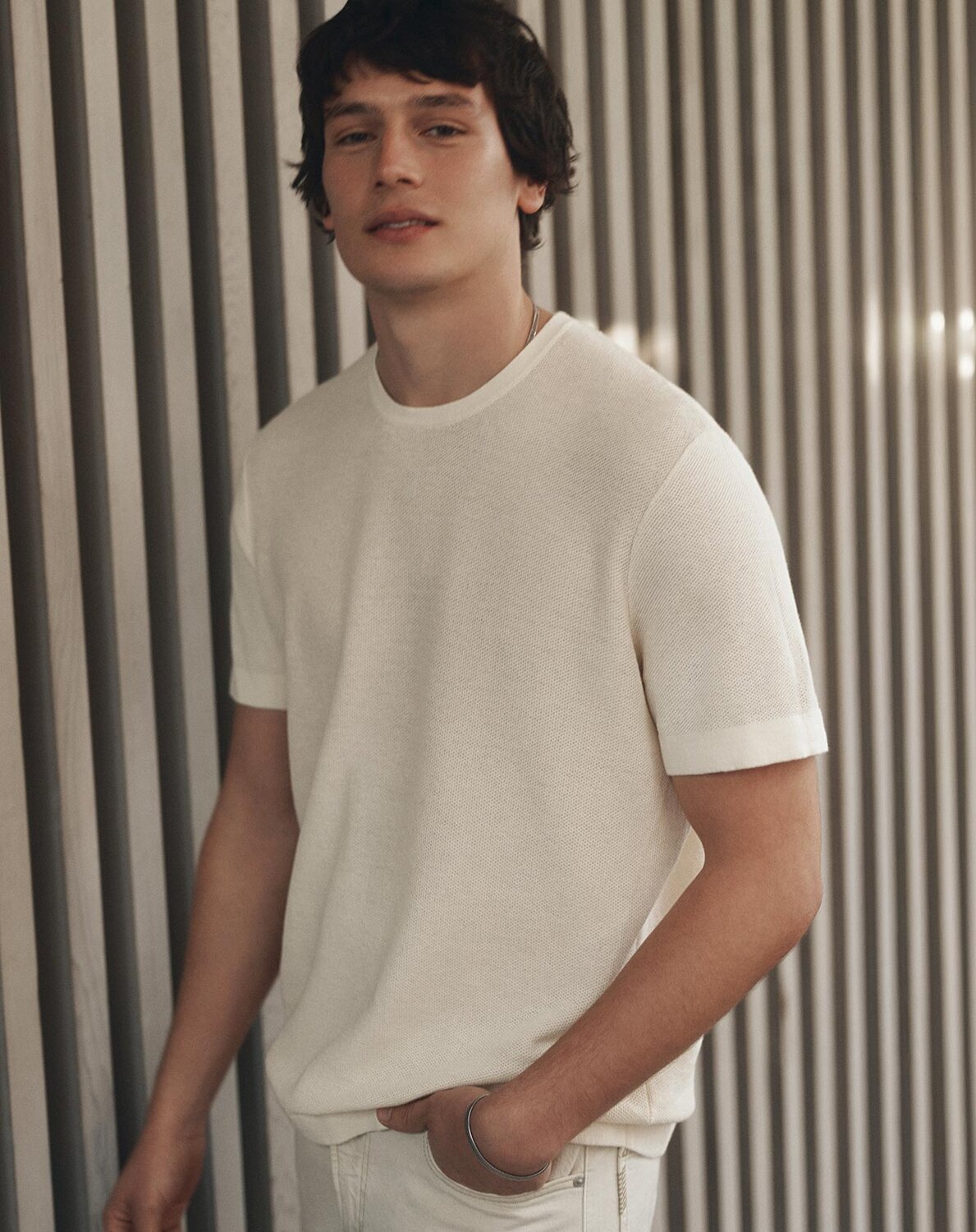 Man wearing a short-sleeve knit shirt and light pants, leaning against a wall with vertical slats.