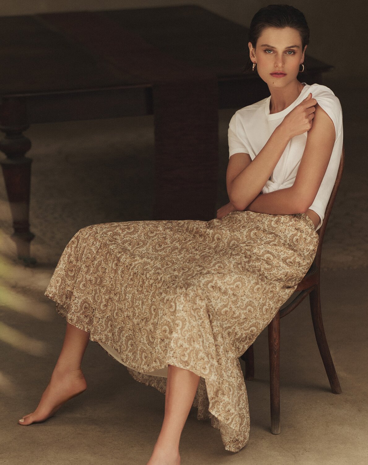 Model seated in a wooden chair wearing a white knotted crewneck tee and a beige paisley-print skirt, accessorized with gold hoop earrings, against a softly lit neutral backdrop.