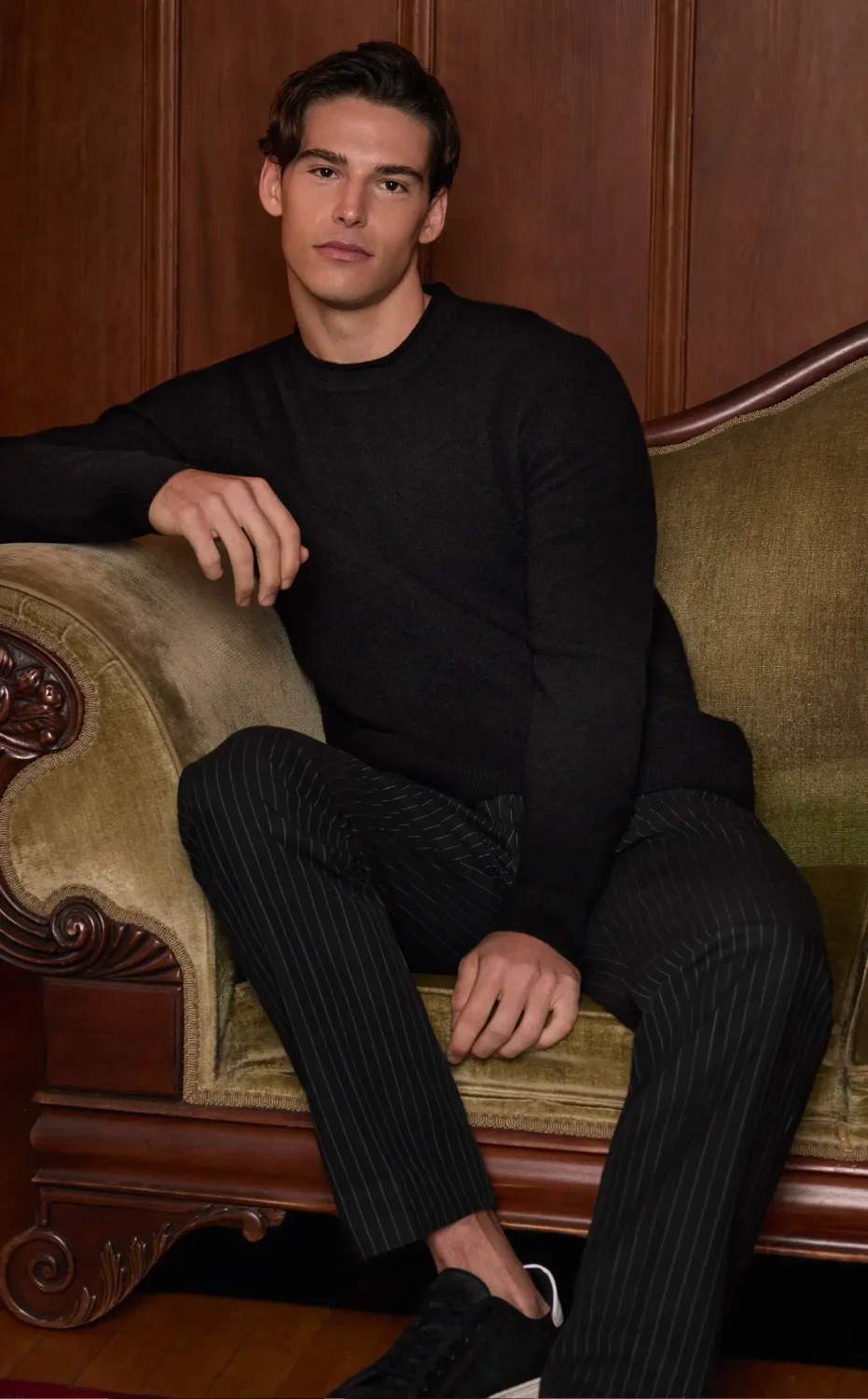 Man sitting on a couch wearing a black crewneck sweater, black pinstripe trousers and black sneakers