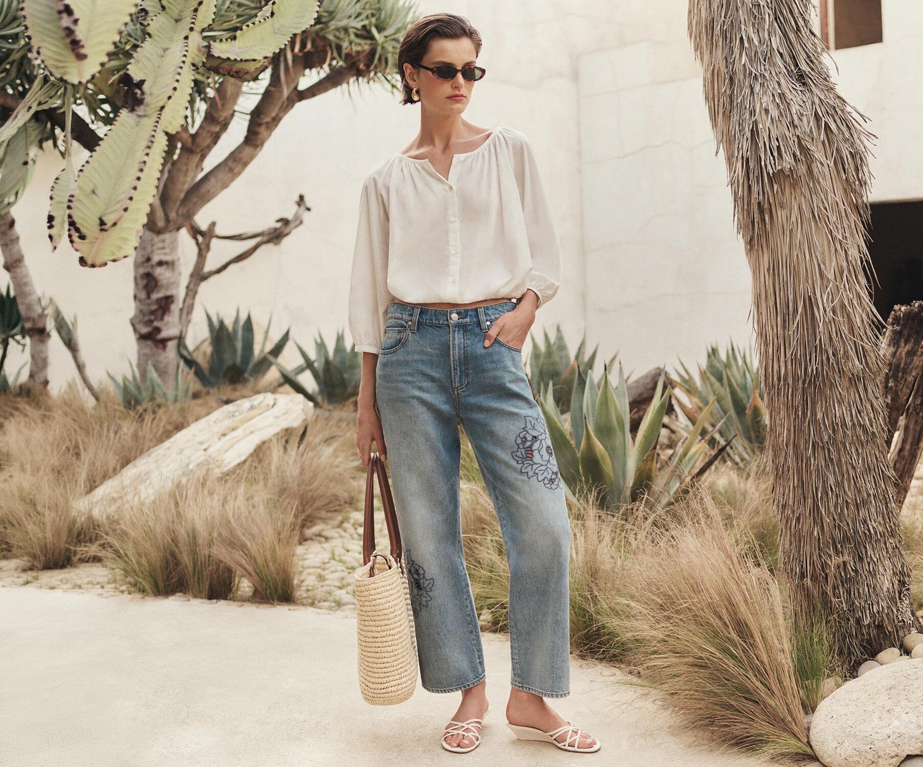 Model wearing a white button-front blouse and light-wash cropped jeans with floral embroidery, accessorized with sunglasses, woven tote, and white sandals, standing in a desert-inspired landscape with cacti, palms, and natural stone accents.