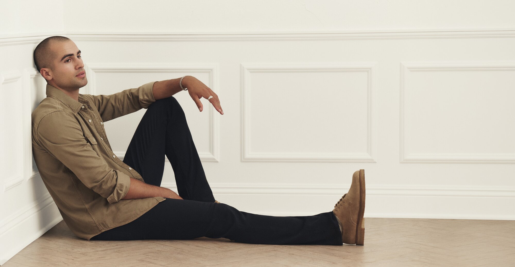 Male model wearing dark wash PAIGE jeans styled with a tan button-down shirt and brown suede shoes.