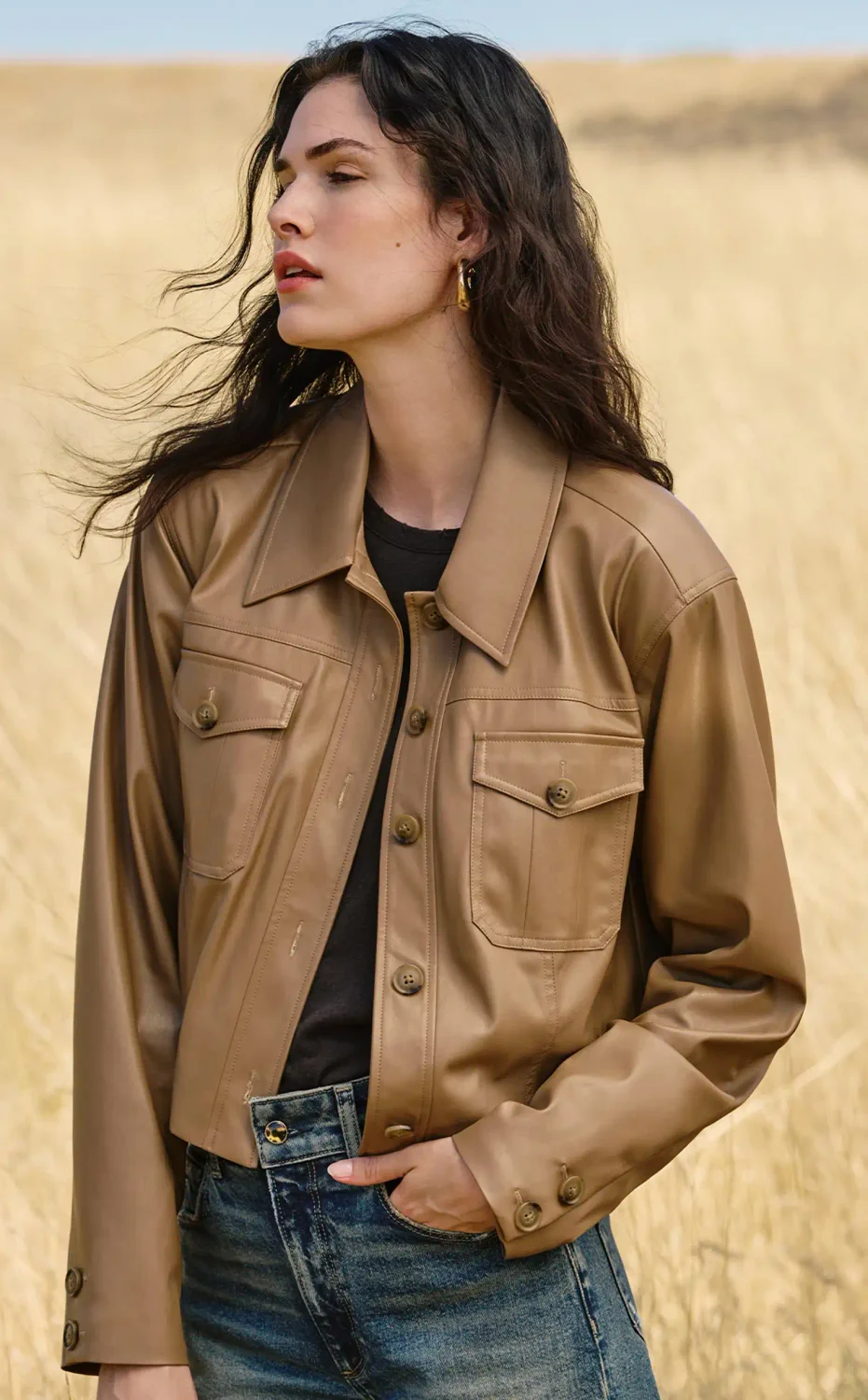 Woman standing outside in a field wearing a light brown faux leather button up jacket, a black top and dark wash jeans