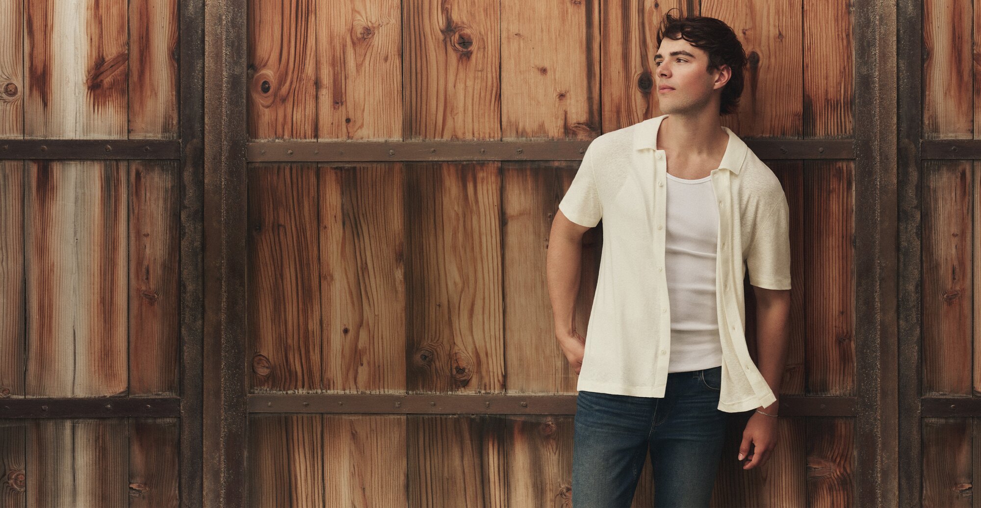 Male model wearing a white short-sleeve button-front shirt layered over a white tank top and dark-wash jeans, standing against a rustic wood-paneled wall.