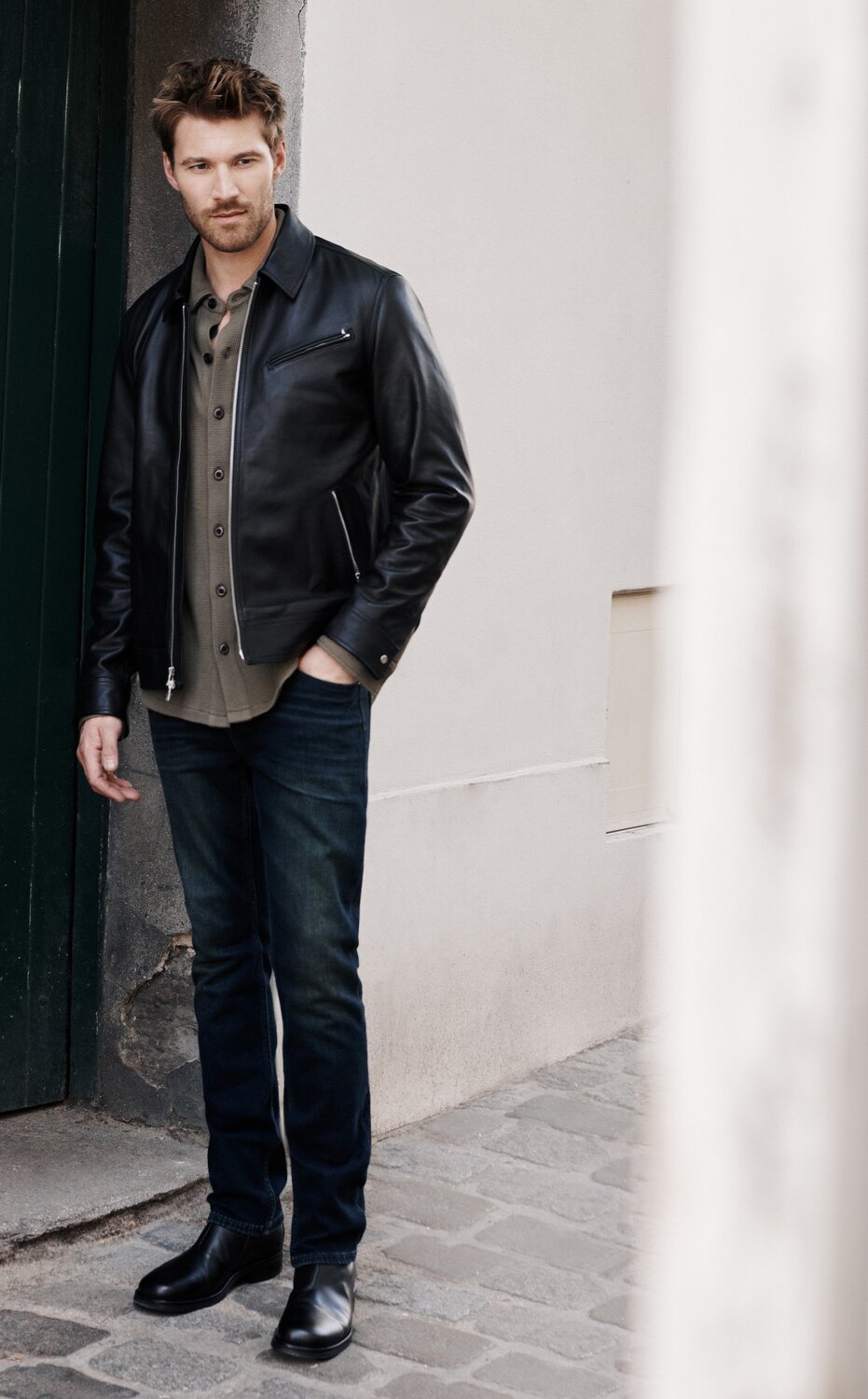 Man wears a black leather moto jacket over a muted green button up and dark wash jeans with black leather boots as he walks the streets of Paris.