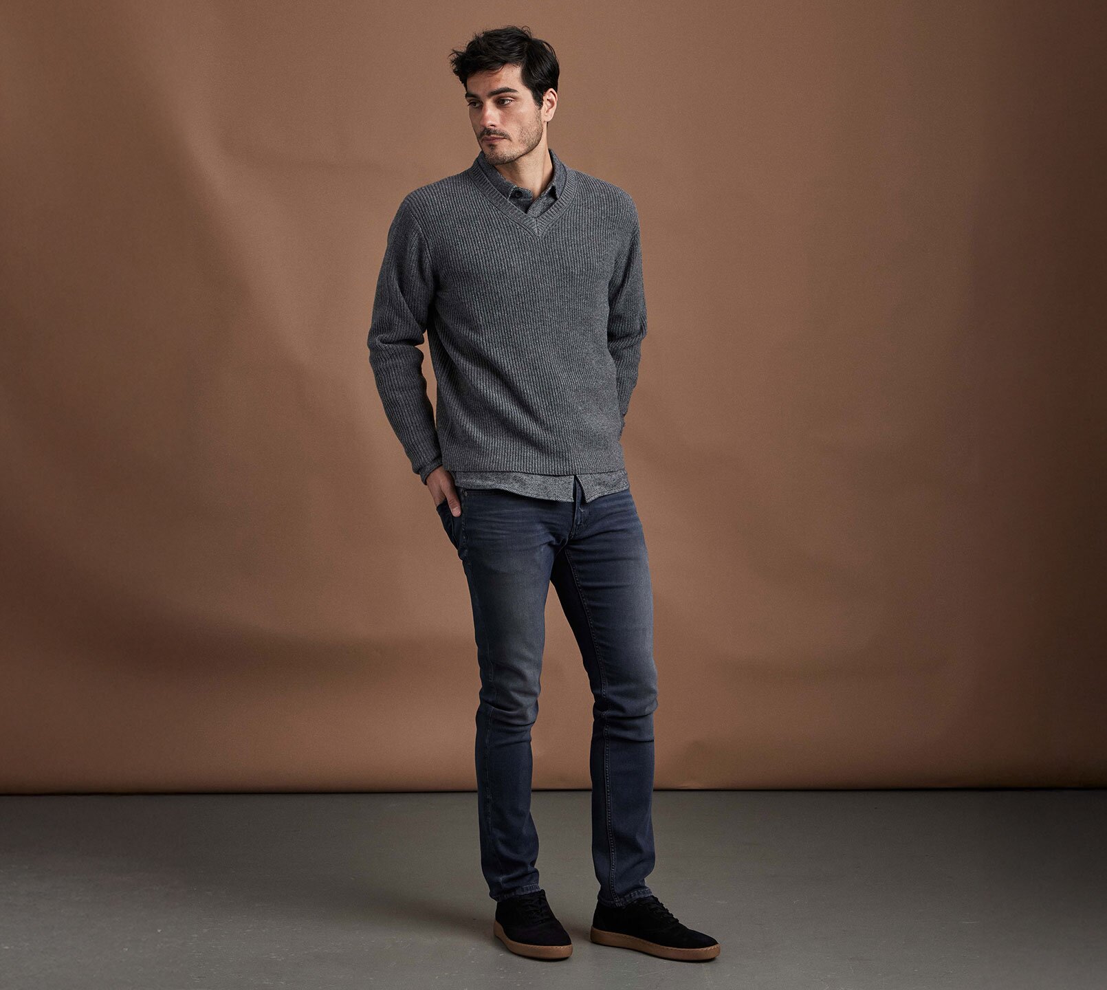 Man standing wearing a grey v-neck sweater over a grey button up shirt, dark wash slim fit jeans and black sneakers