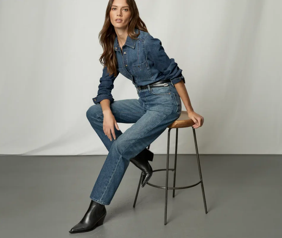 Woman sitting on a stool wearing a dark wash button up denim shirt, dark wash straight leg jeans, black boots, and a black belt