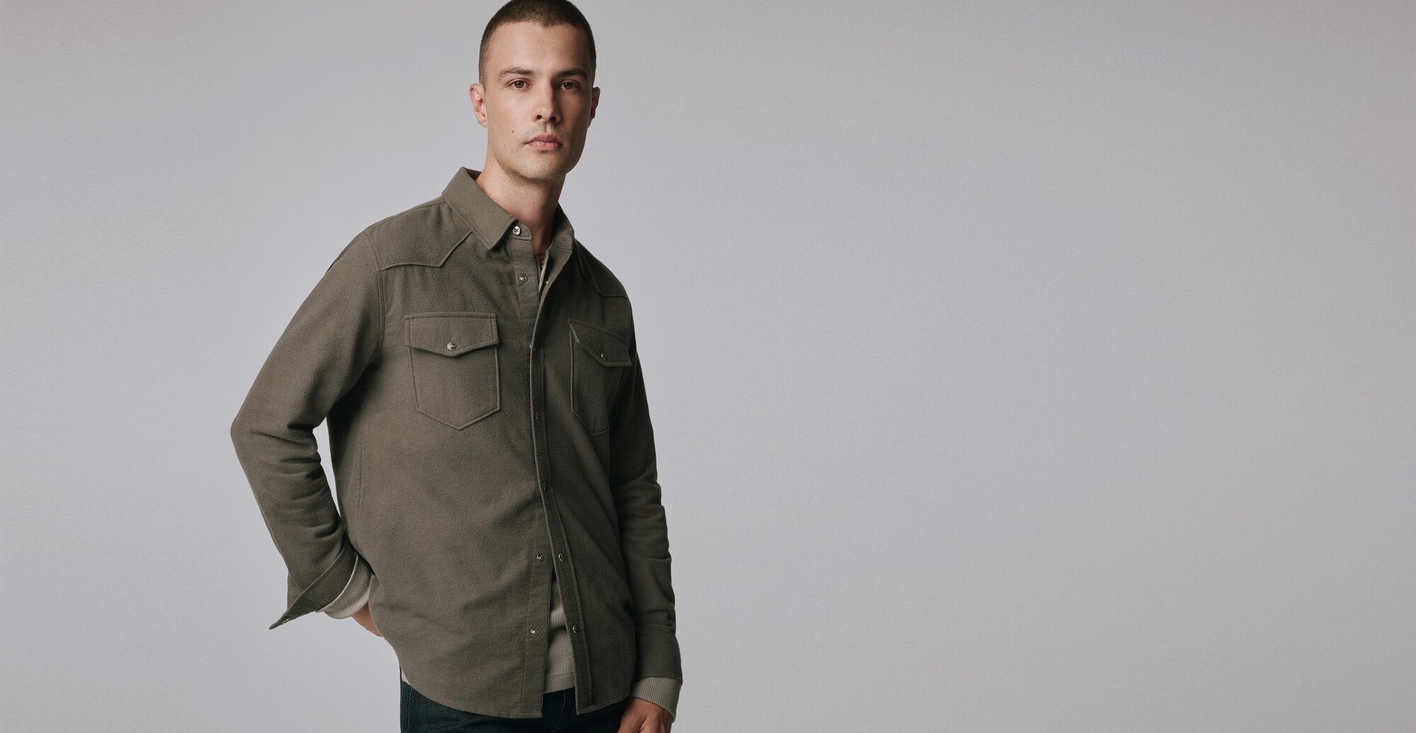 Man wearing a grey button-up shirt over a grey long sleeve tee and dark wash jeans