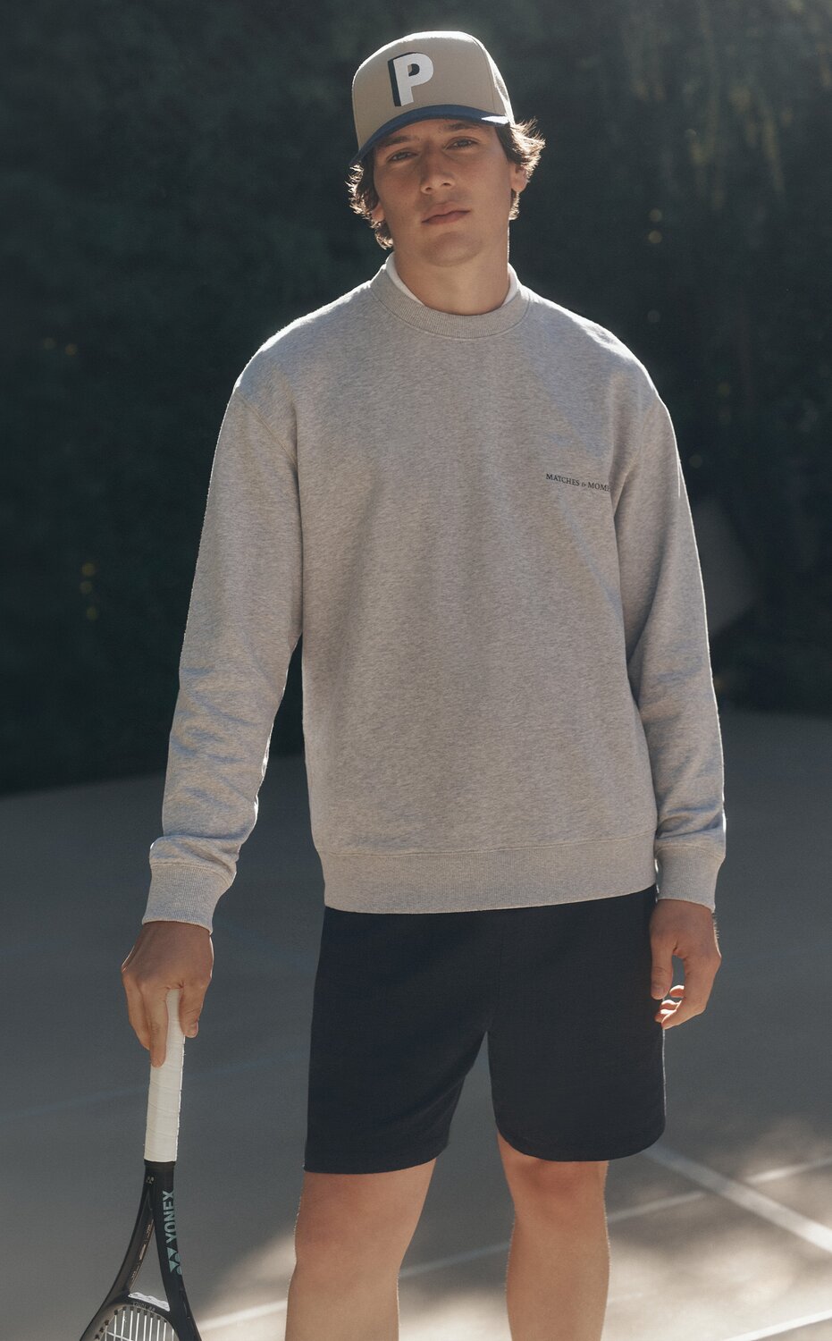 Man wearing a grey pullover with a pair of navy shorts and a cap decorated with the letter P, posed on the tennis court with a racket in hand.