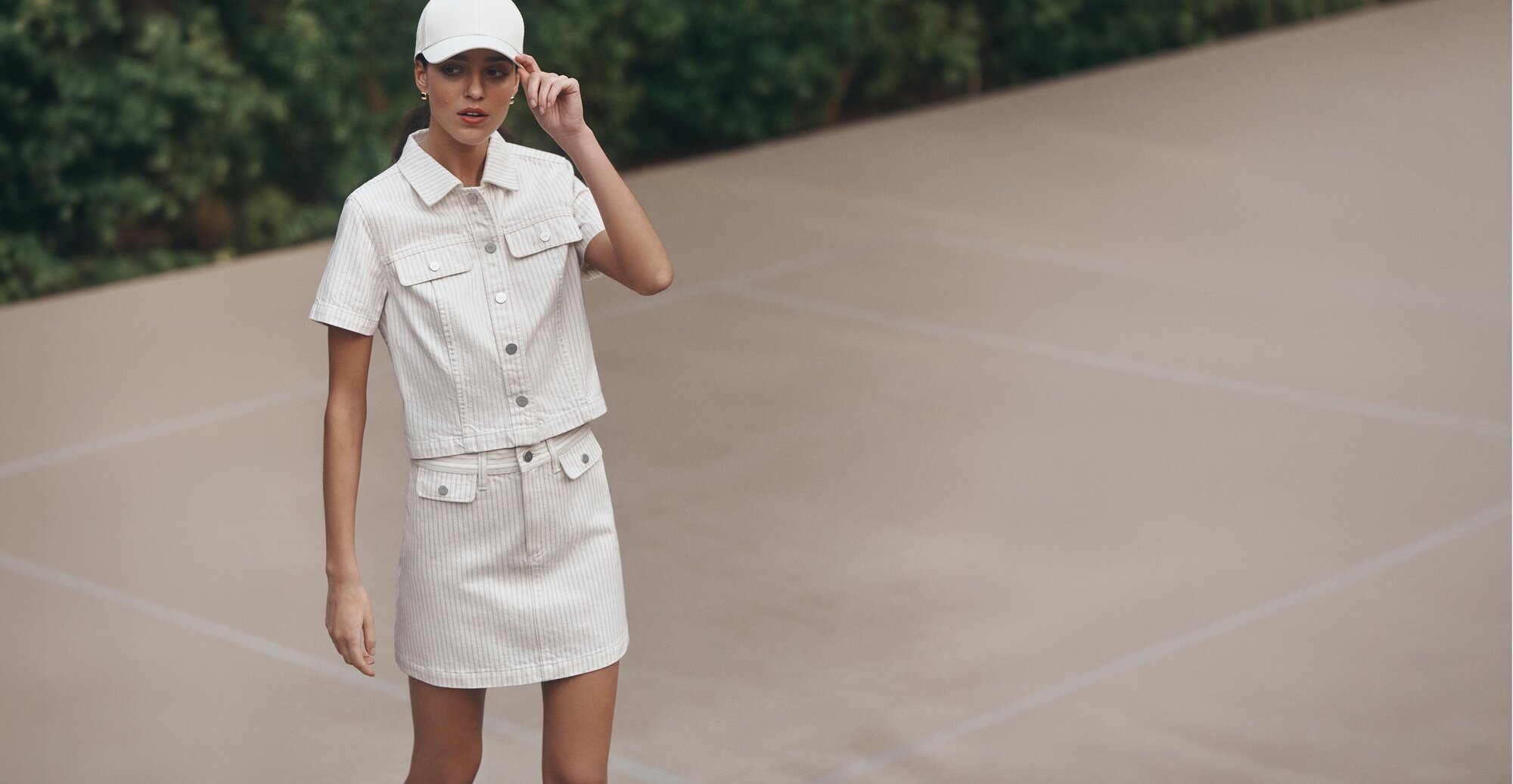 Woman posed at a tennis court wearing a white striped denim matching top and skirt with a white cap.