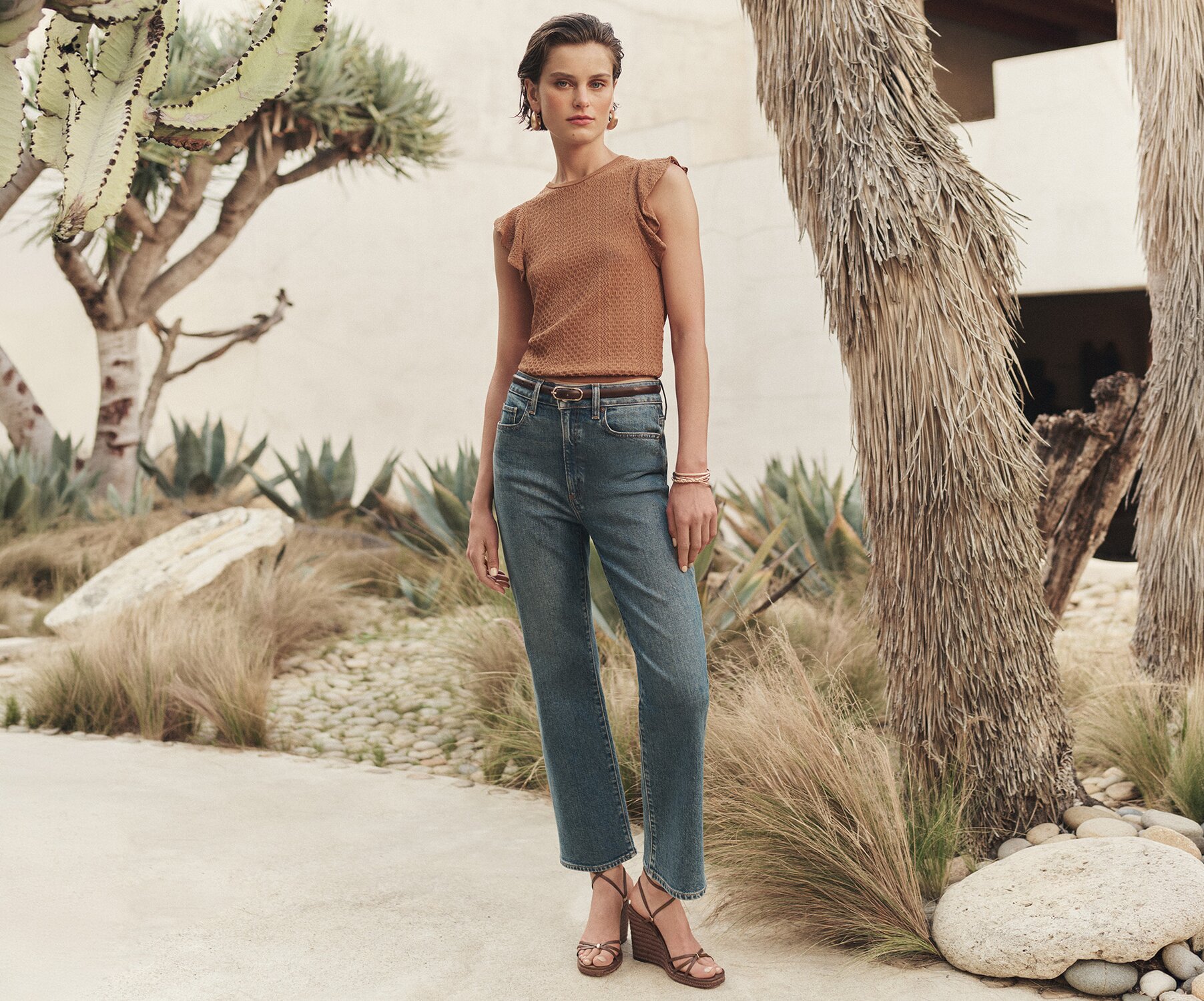 Woman wearing an orange flutter-sleeve knit top with a pair of medium wash high waisted straight-leg jeans and strappy brown wedge sandals, posed in a courtyard surrounded by palm trees.