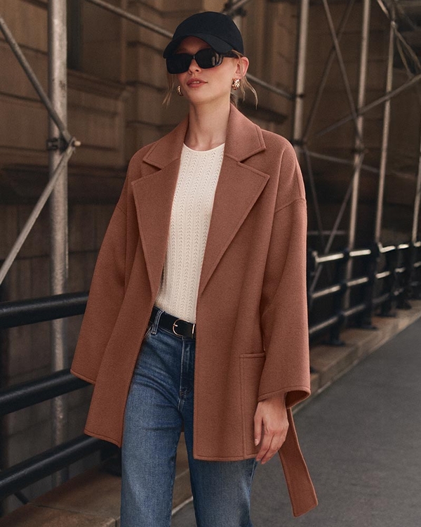 Woman wearing camel brown robe coat over a white top with medium wash jeans, a black belt, black boots, black baseball cap, and black sunglasses