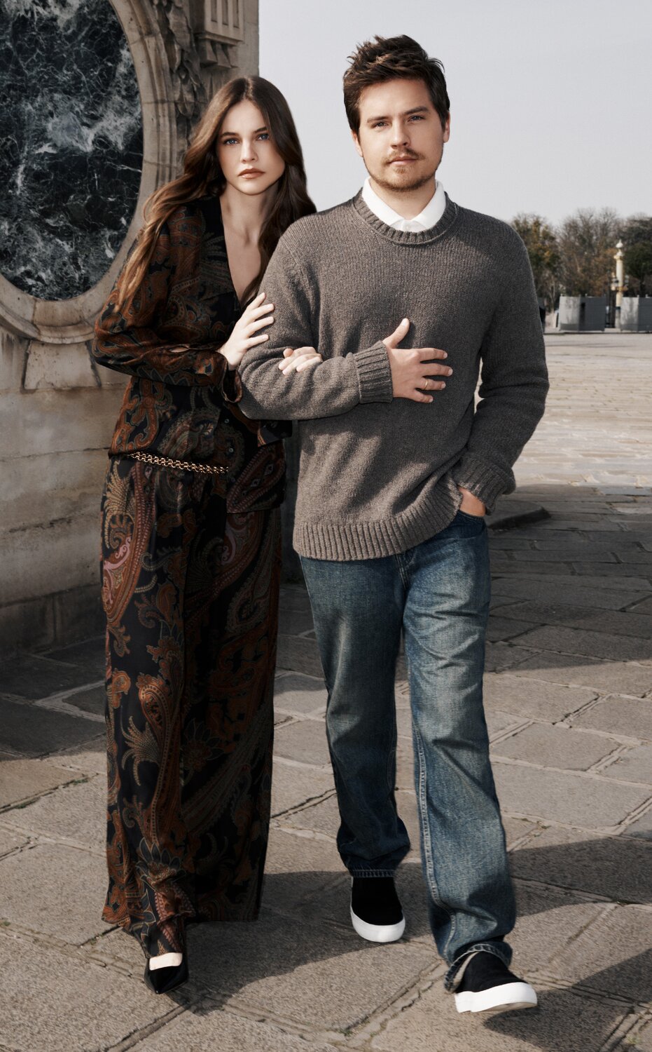 Barbara Palvin wears a dark brown paisley print top and pant set with a gold chain belt as she walks arm-in-arm with Dylan Sprouse, who wears a grey crew neck sweater over a white button-up and a pair of medium wash jeans.