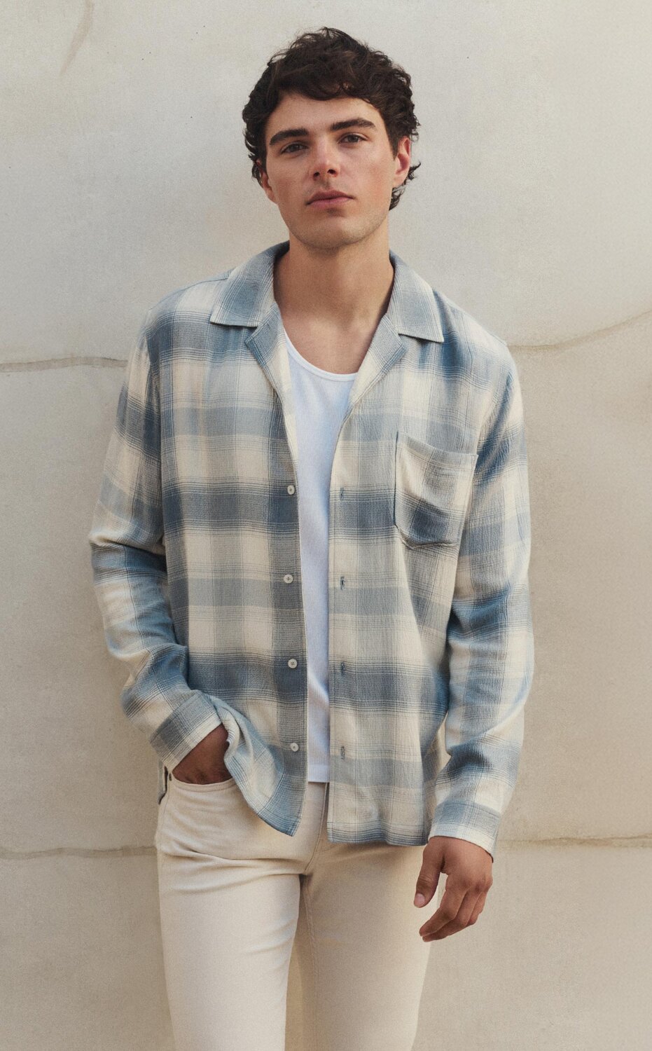 Male model wearing a light blue and cream plaid flannel long-sleeve button-up shirt over a white ribbed tank with light cream colored denim jeans, all by PAIGE, standing in front of a light cream colored wall.
