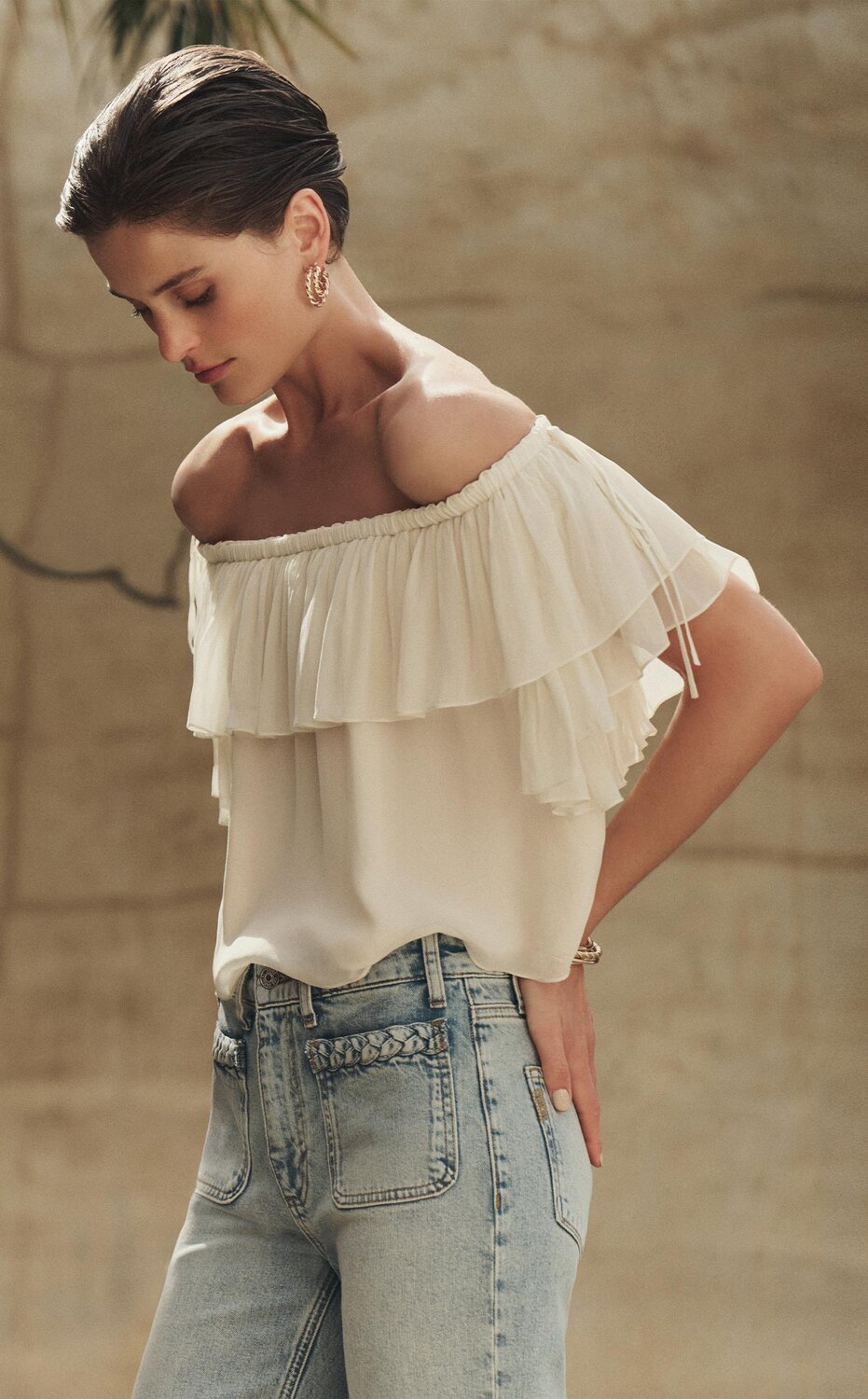 Woman wearing a white off-the-shoulder ruffle blouse and light-wash high-rise jeans with braided pocket detailing, standing against a sunlit stone wall with palm trees in the background.