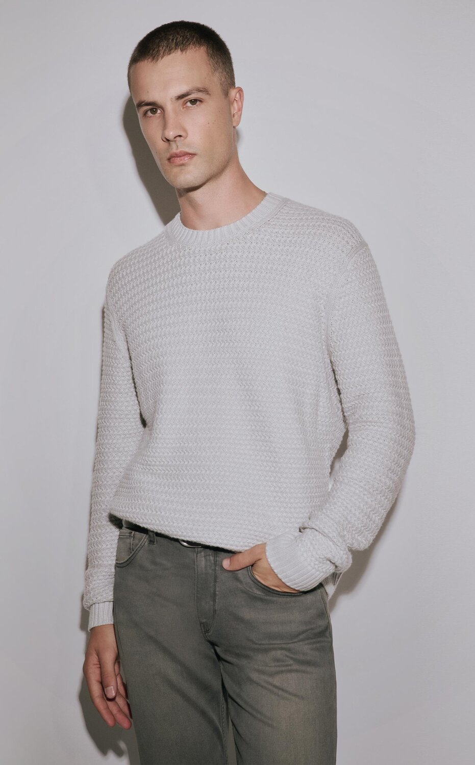 Man wearing a light grey sweater with grey wash jeans and a black belt