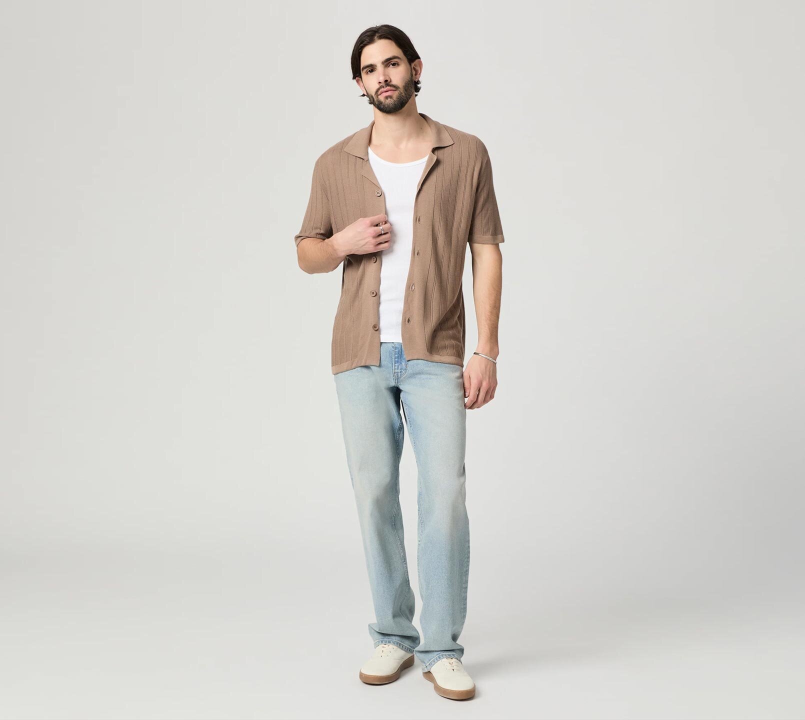 Man wearing a brown short sleeve button up sweater shirt, white tank top, light wash jeans and bone sneakers