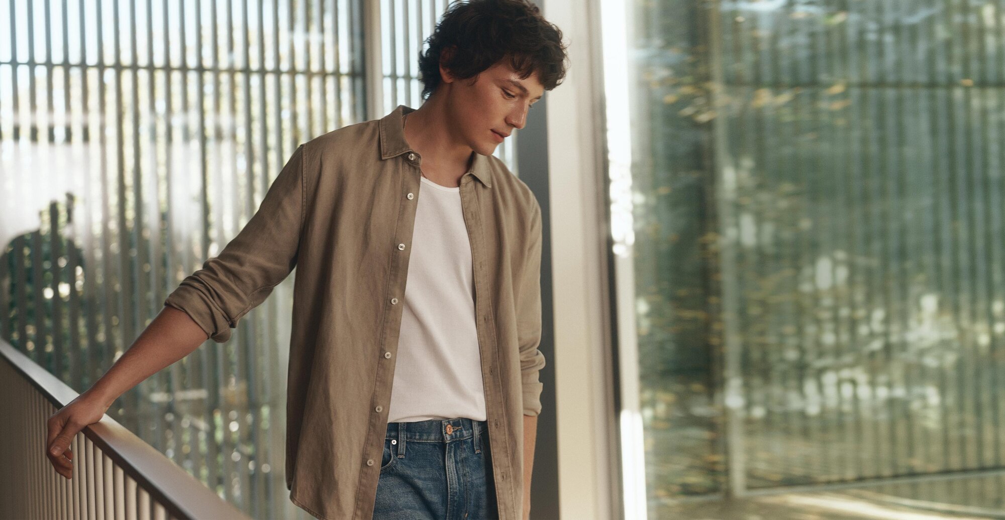 Man wearing a light beige button-down shirt over a white tee and blue jeans looking out a window.