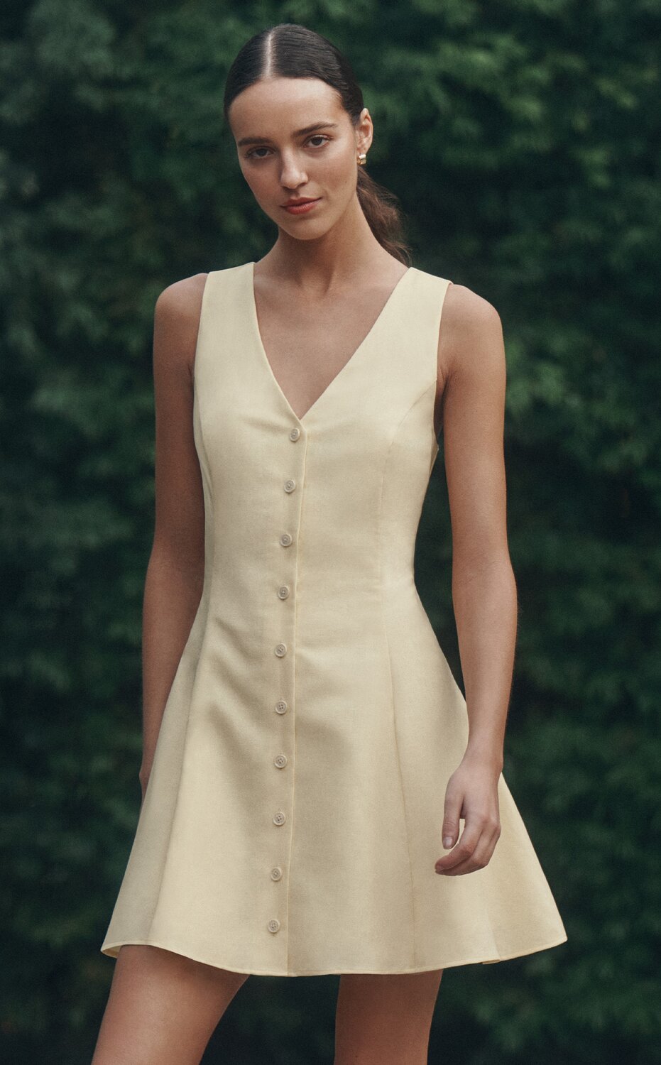 Woman wearing a sleeveless yellow button-front dress standing outdoors against green foliage.