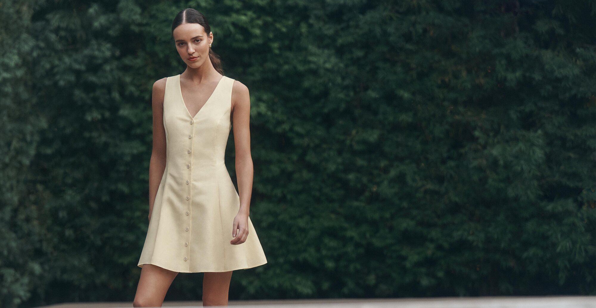 Woman wearing a sleeveless yellow button-front dress standing outdoors against green foliage.